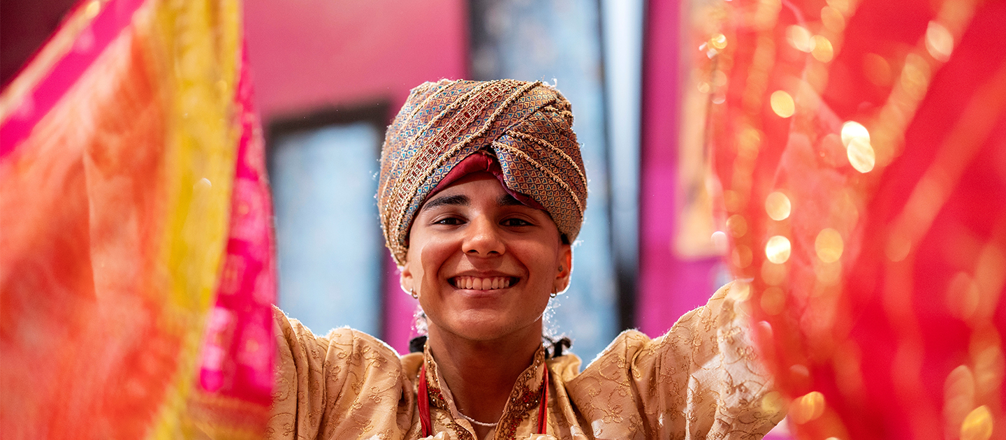 Teenager in a turban smiling into the camera between two folds of bright coloured and sequined cloth.