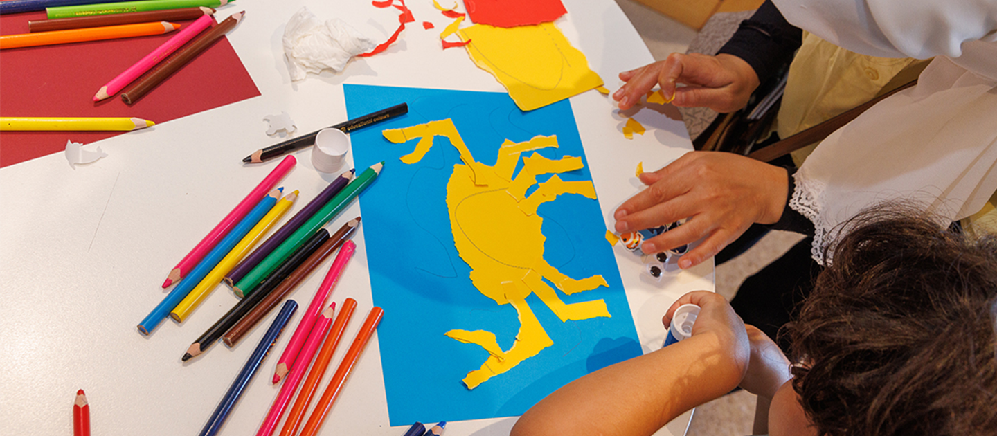 View from above of a craft table with a flat paper crab picture and people decorating it. Surrounded by paper, pencils and other supplies. 