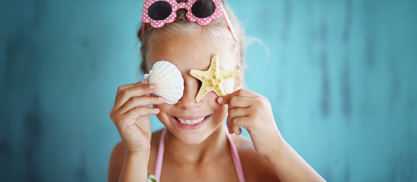 Child wearing sunglasses on head and beachwear holding up two different shaped shells over her eyes. 