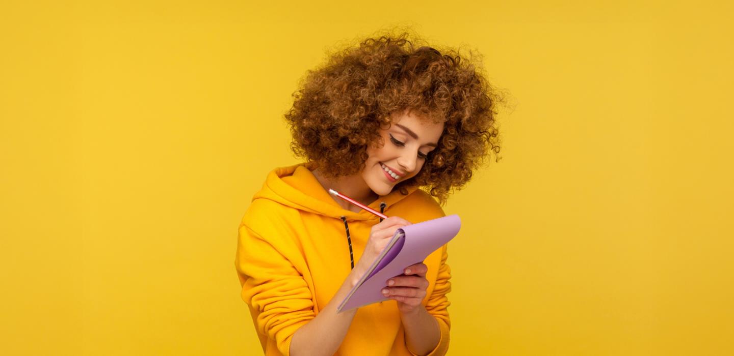 Young woman with curly hair and wearing a yellow hoodie writing on a notepad in front of yellow background.