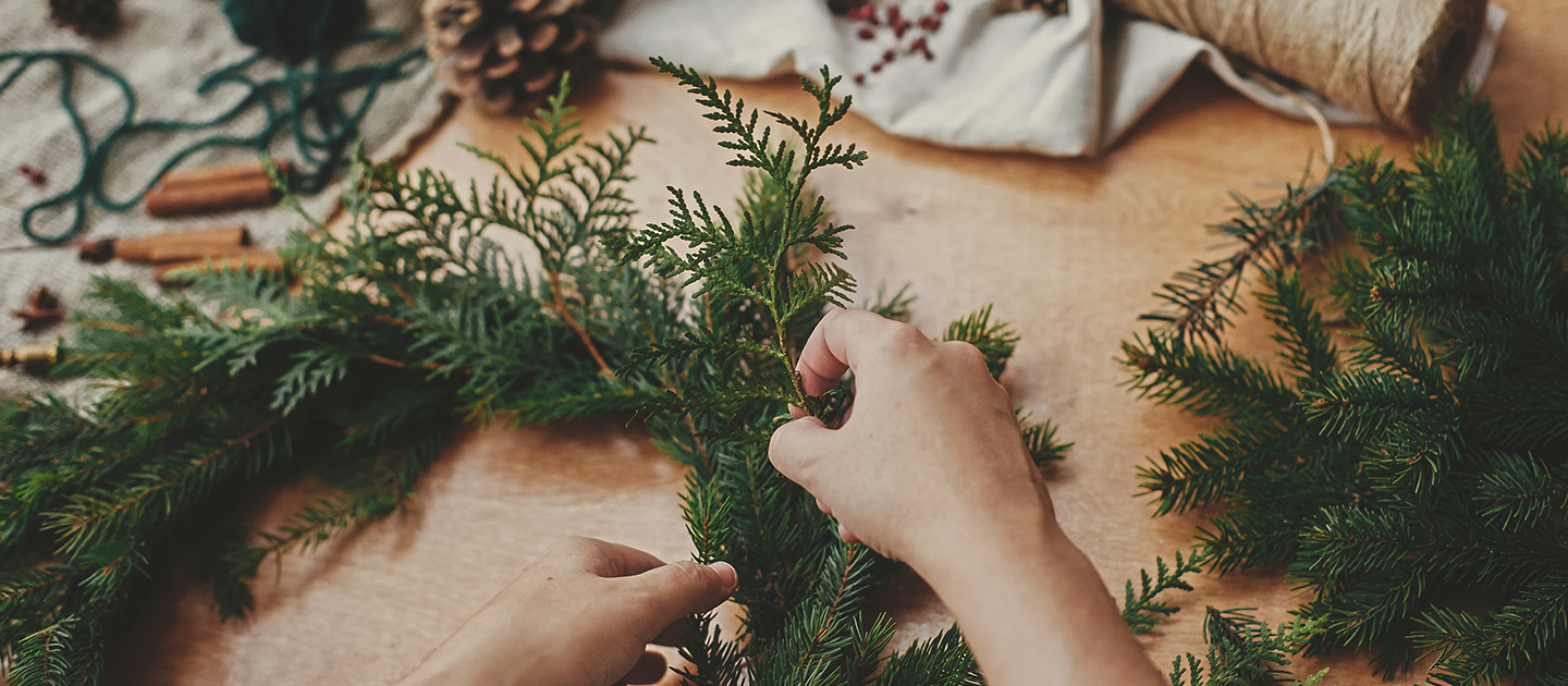 Close up of hands crafting a Christmas wreath from green foliage with pinecones and string in the background. 
