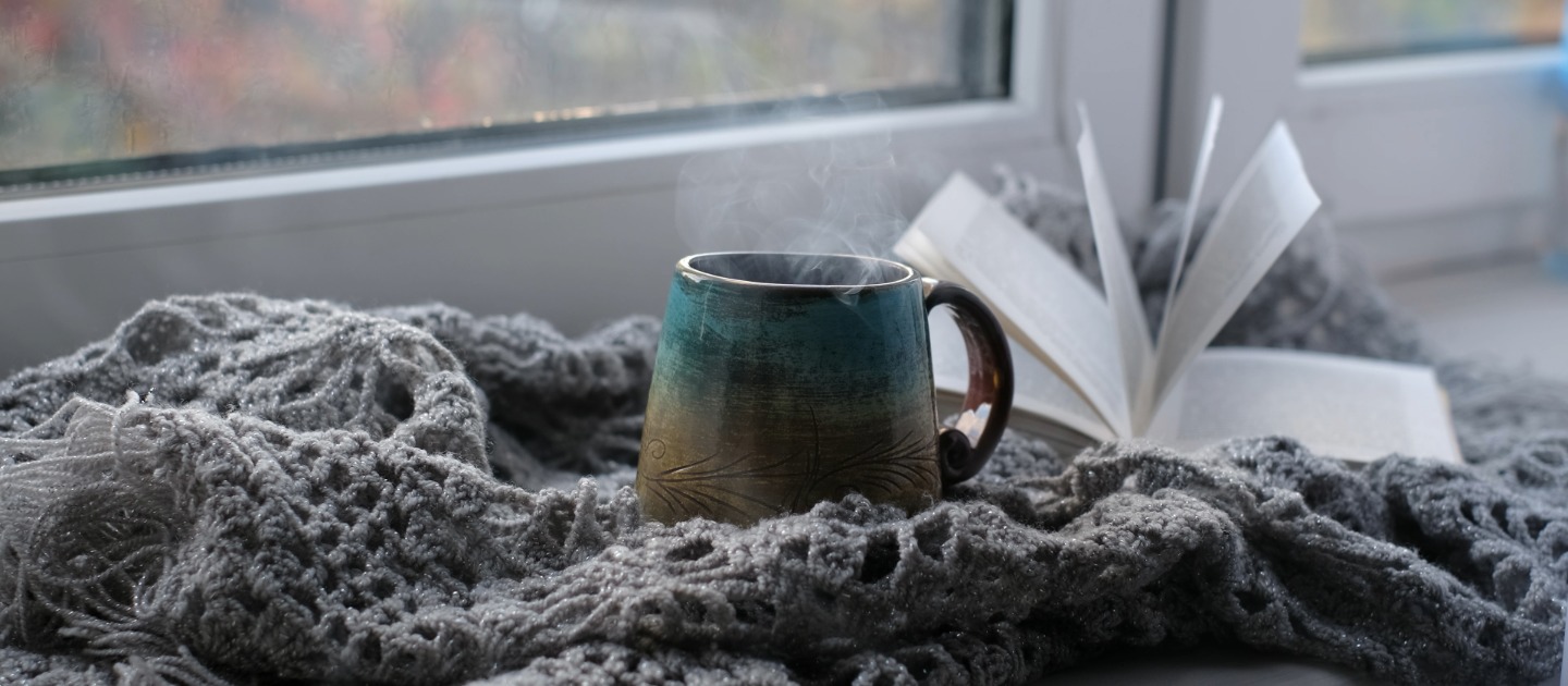 Steaming hot drink and book, resting on a grey blanket by a window with winter weather outside. 
