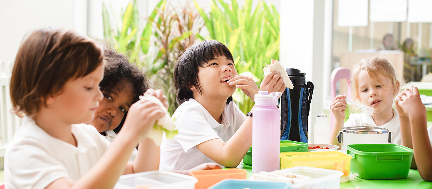 A group of children sat around a table eating a healthy packed lunch.