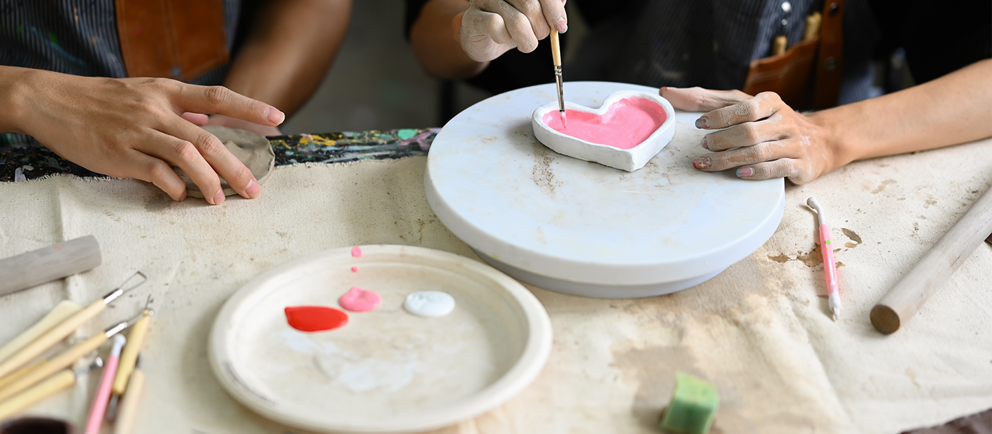 View of a worktable with two young people decorating an air clay heart-shaped dish.
